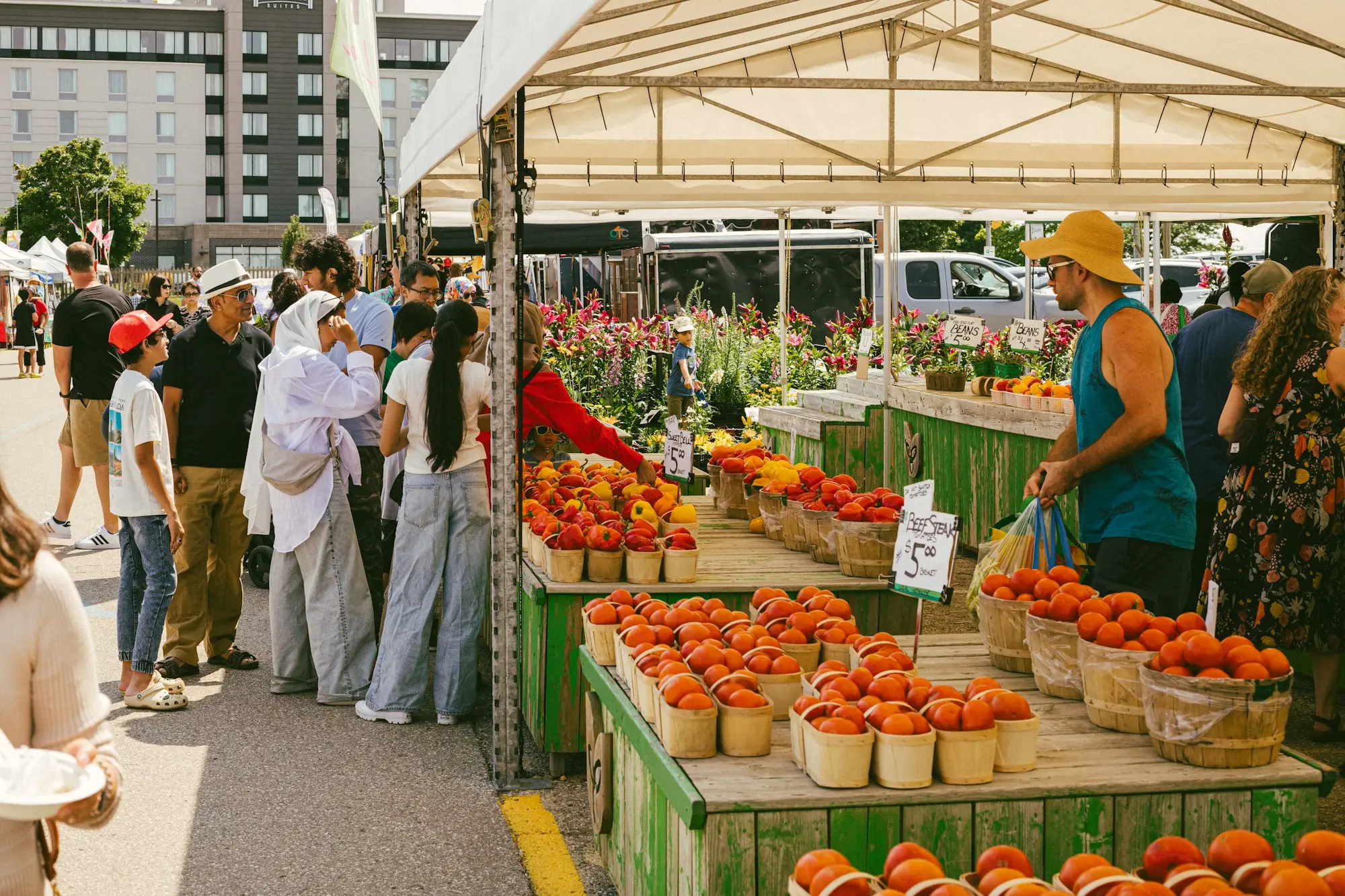 Shoppers browse baskets of tomatoes and sweet peppers under a canopy at a sunny outdoor farmers market